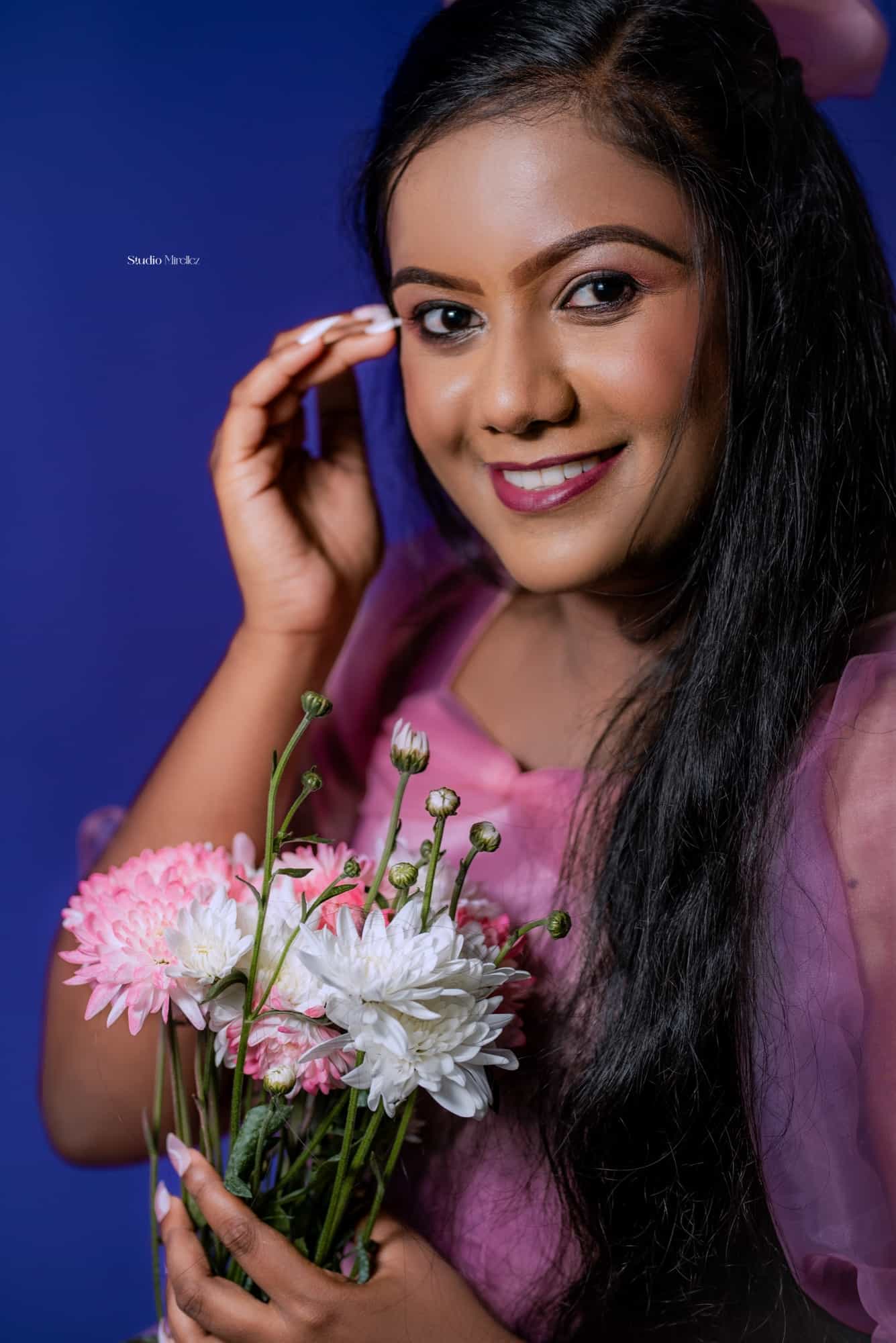 Close-up portrait of Githmini smiling against a deep violet backdrop, holding a bouquet of pink and white chrysanthemums