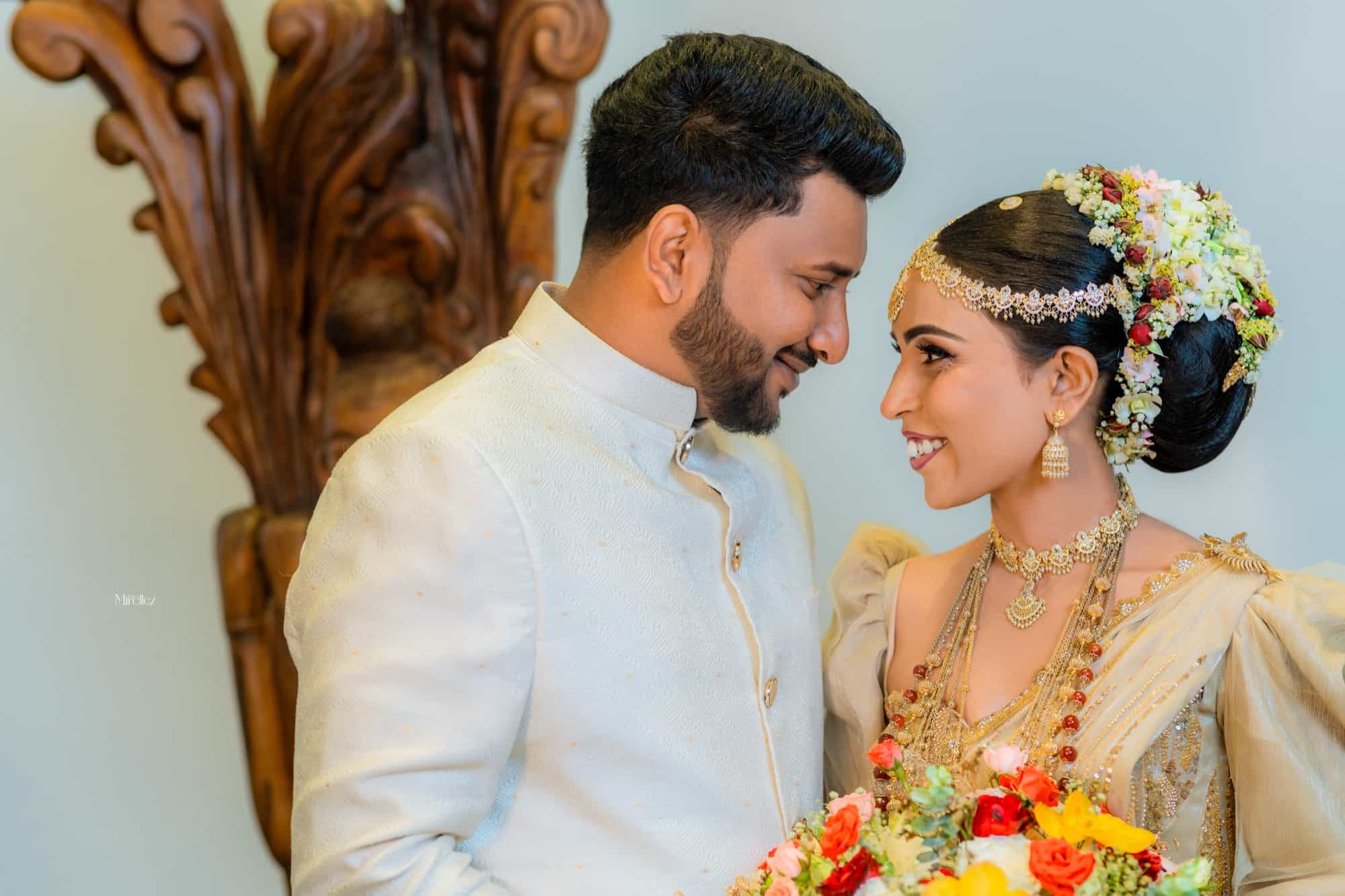 Close profile of the couple smiling at each other, the bride's floral-wreathed bun and layered gold necklaces catching the light