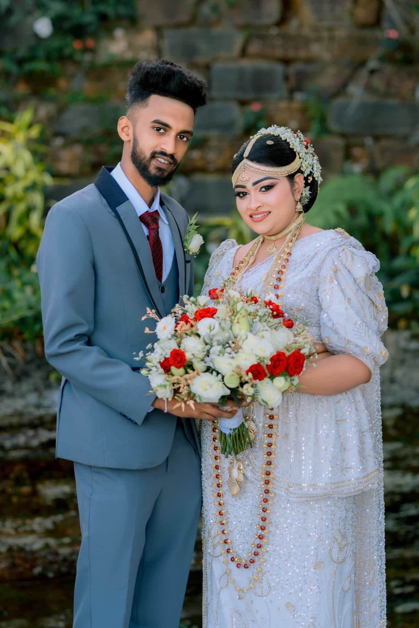 Bride and groom against a stone wall framed by ferns, she in a beaded white Kandyan saree, he in a slate-blue three-piece suit