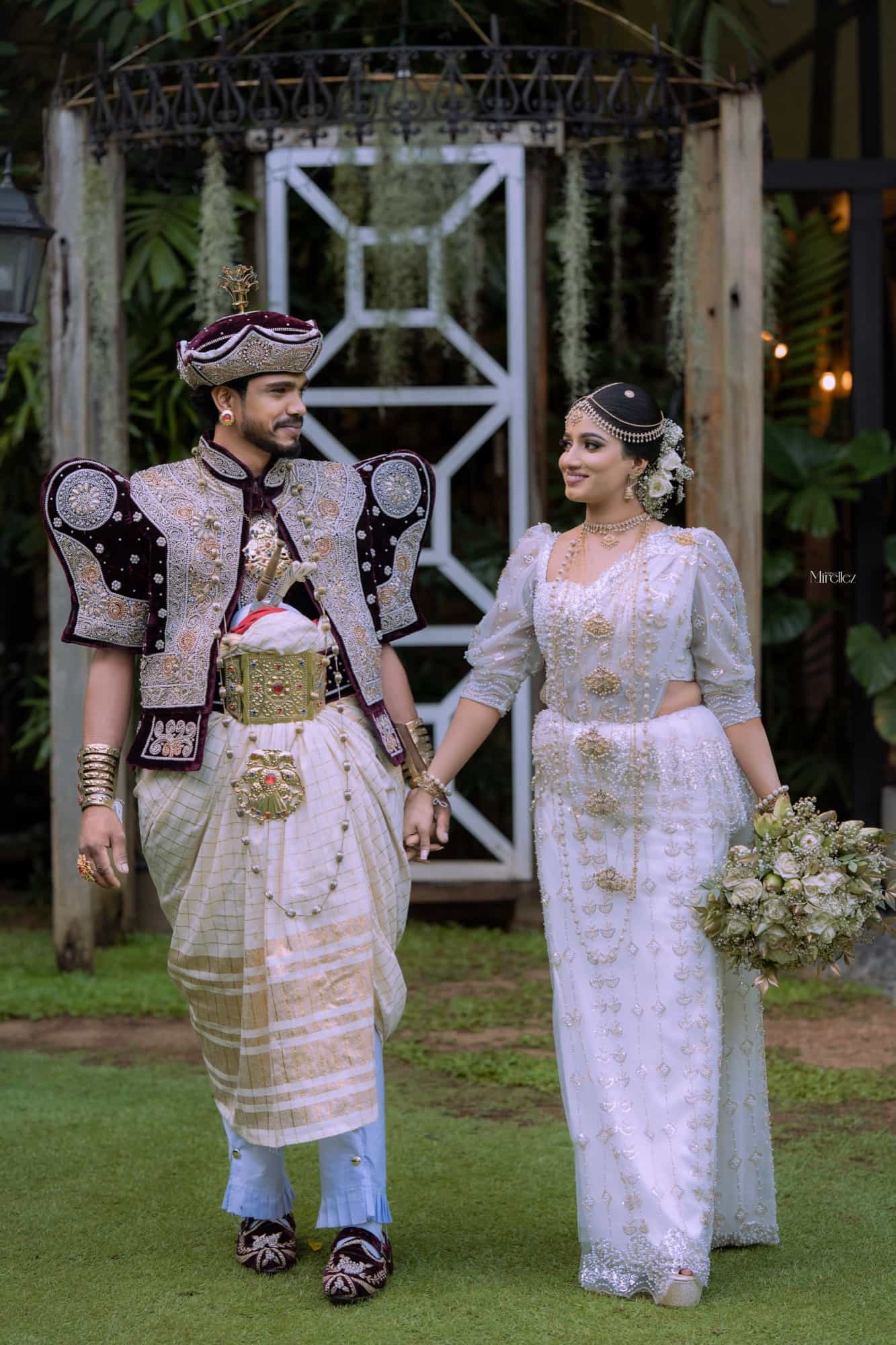 Full-length portrait of the couple holding hands on a lawn before a white wooden arch draped with trailing greenery