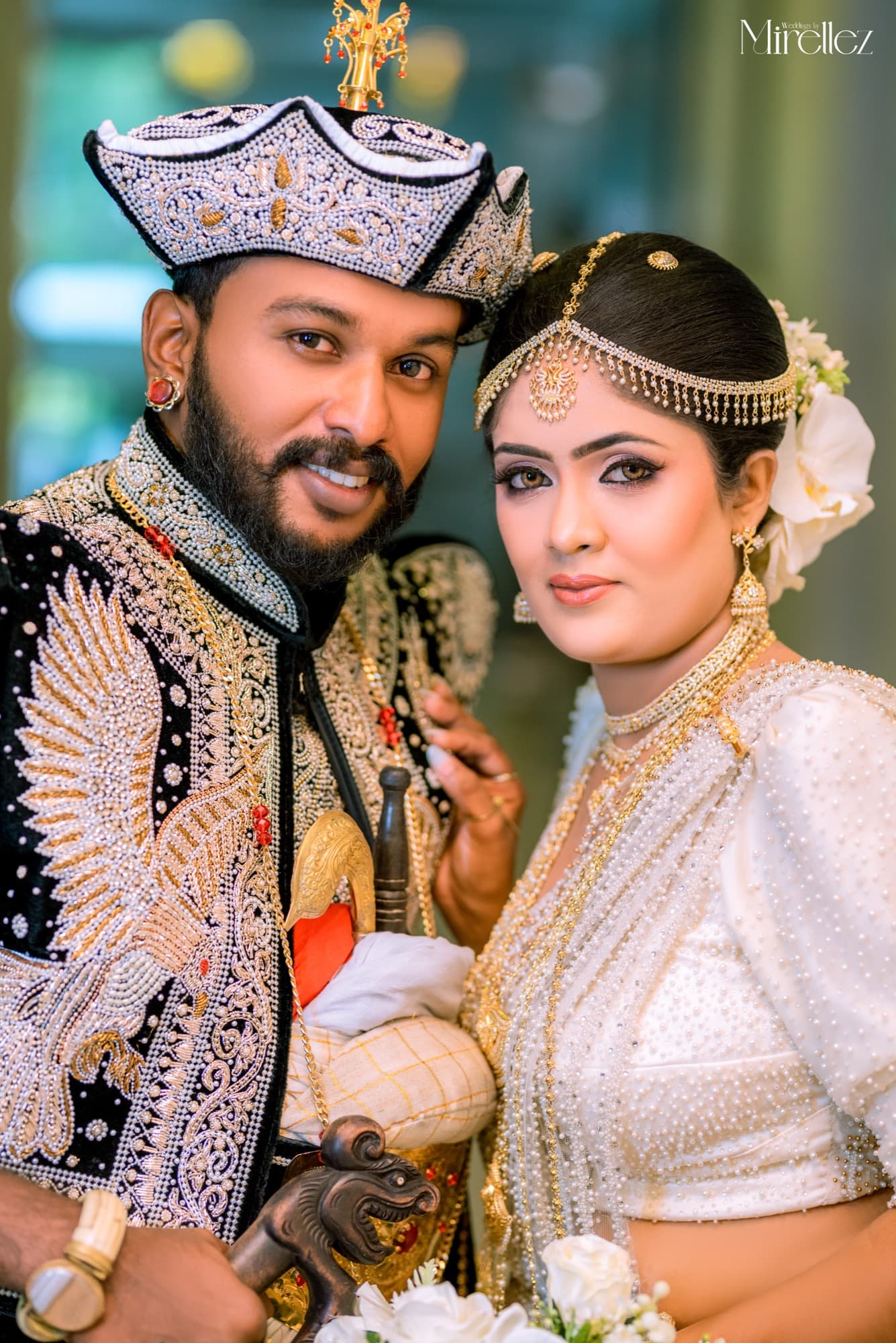Close-up portrait of a Kandyan bride and groom, he in a jewelled nilame headdress and pearl-embroidered black jacket, she in a white pearl osari with a gold headpiece