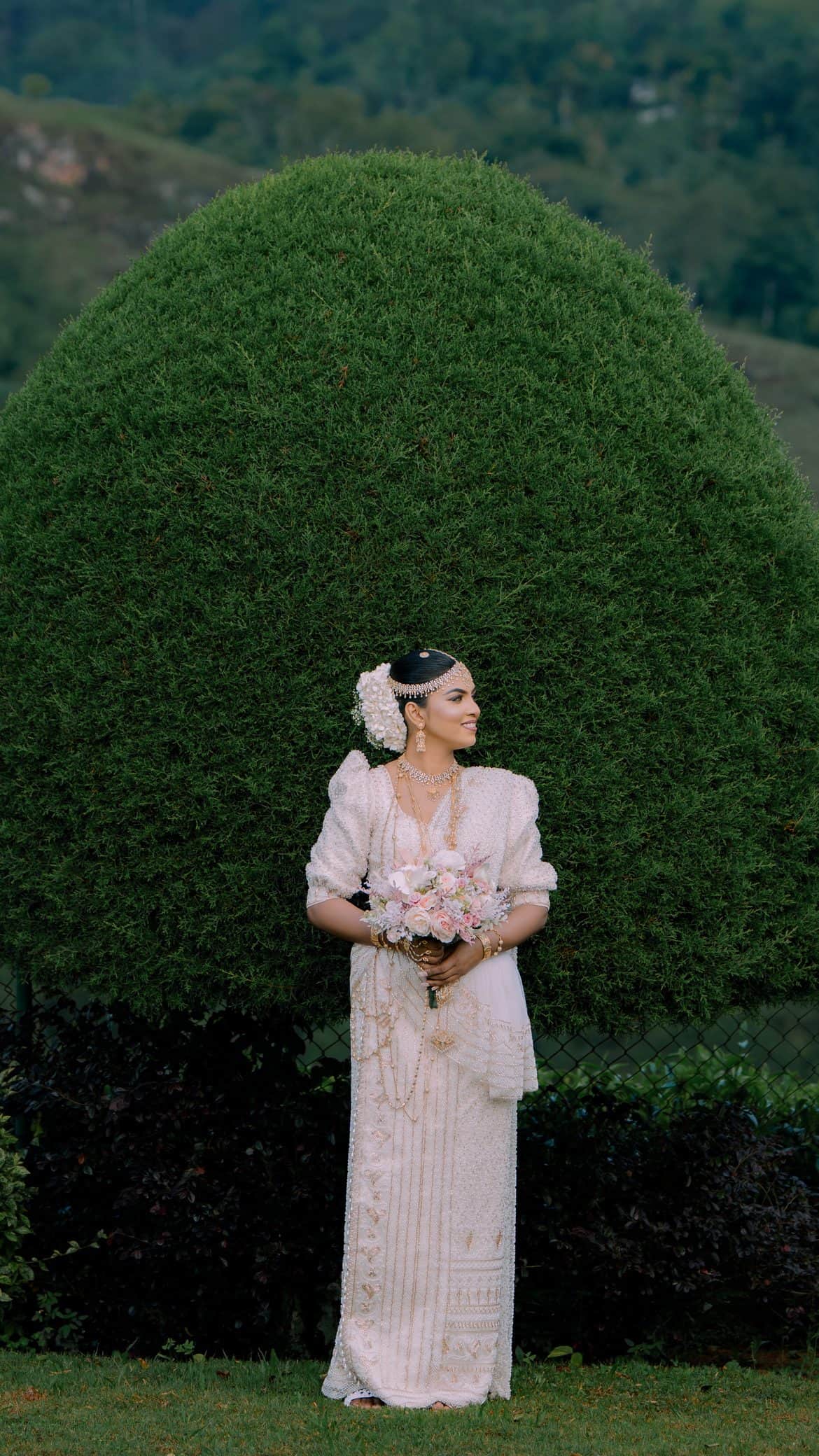 A bride in a pearl-beaded osariya standing before a rounded topiary, misty hill country behind