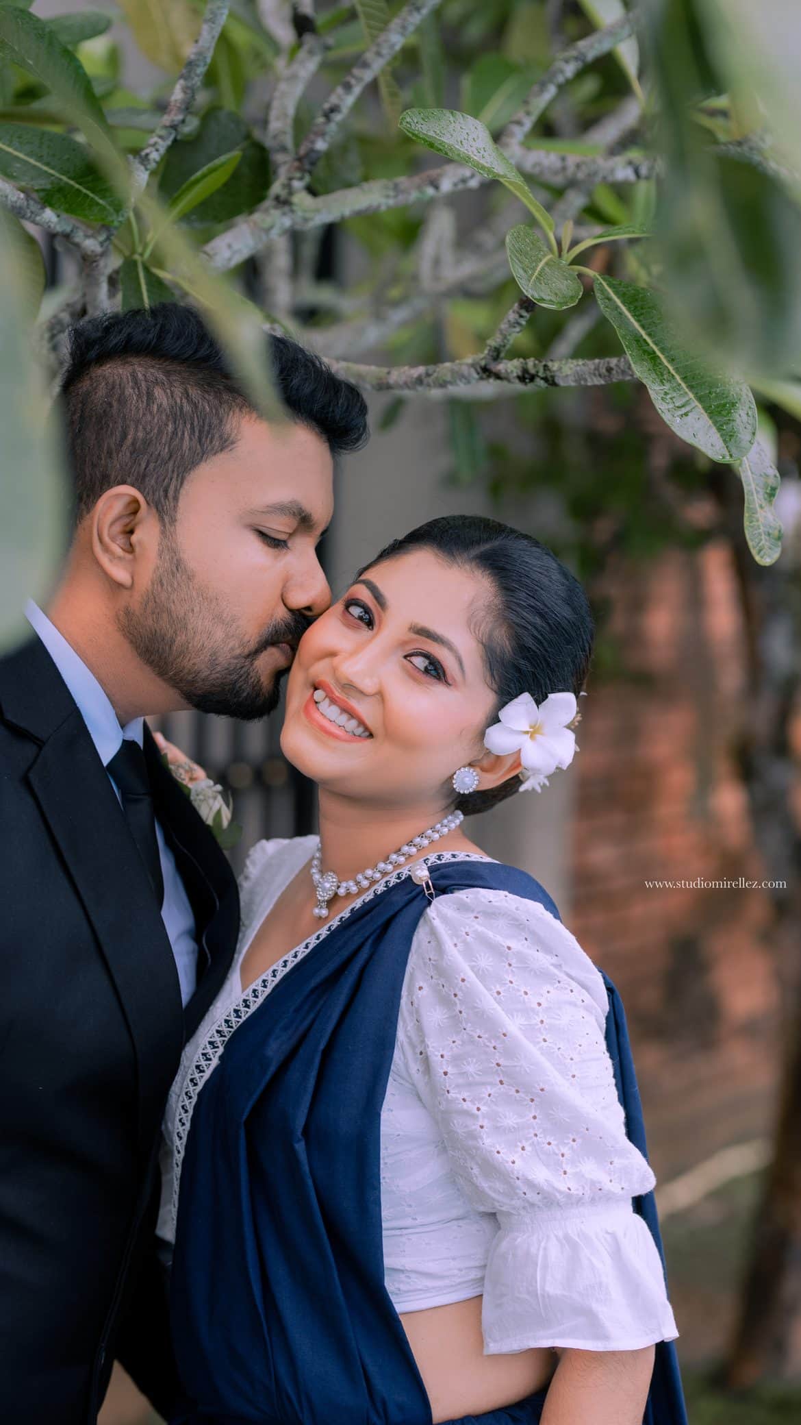 A groom kissing his fiancee's cheek beneath a frangipani tree, a white blossom in her hair