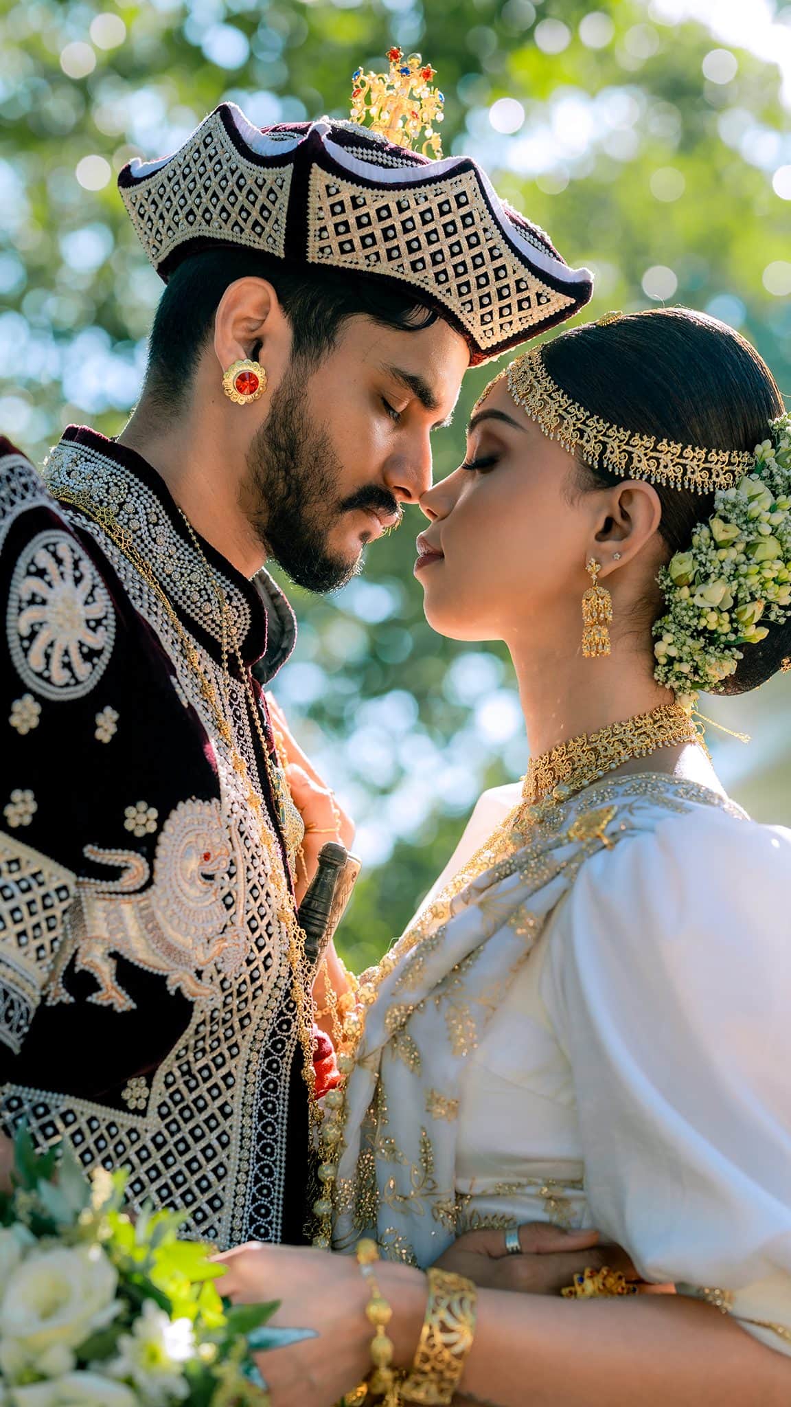 A Kandyan groom in a jewelled crown and bride in gold headdress, nose to nose in dappled light